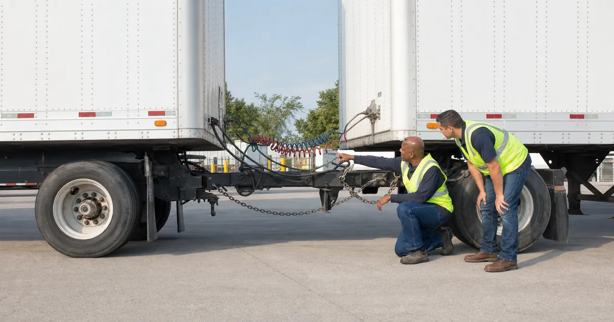 CDL instructor and trainee inspecting converter dolly air lines, safety chains, and coupling hardware between two commercial trailers